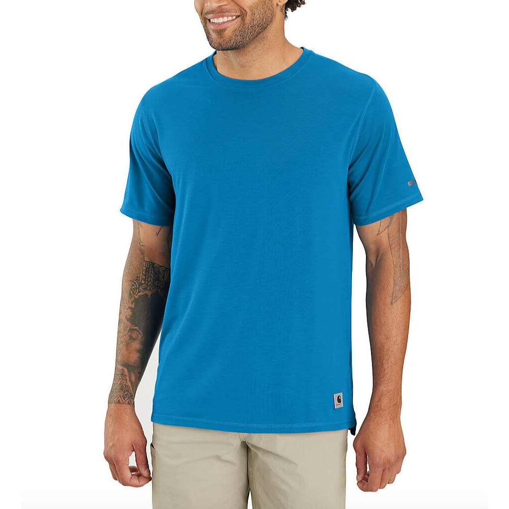 A man is wearing a bright blue short-sleeve T-shirt with a small logo tag, paired with beige pants. He has tattoos on his arms and a smile on his face.