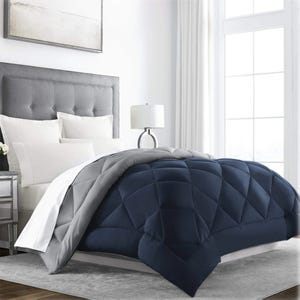 A down-alternative reversible comforter with a navy and gray color scheme, featuring a quilted design. It is displayed on a bed with a gray tufted headboard, accompanied by white pillows and a side table lamp in a bright, modern bedroom.