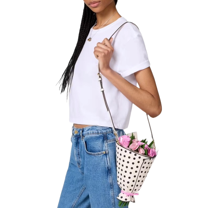 The Kate Spade Flower Bouquet Crossbody is a polka dot bag shaped like a flower bouquet, complete with pink flowers peeking out. It's worn over the shoulder with a leather strap, paired with a casual white t-shirt and jeans.