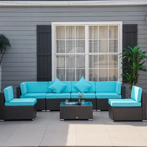 Outdoor patio furniture set featuring a modular black wicker sectional with turquoise cushions and a rectangular glass-top coffee table, set against a gray house with a window and plants.