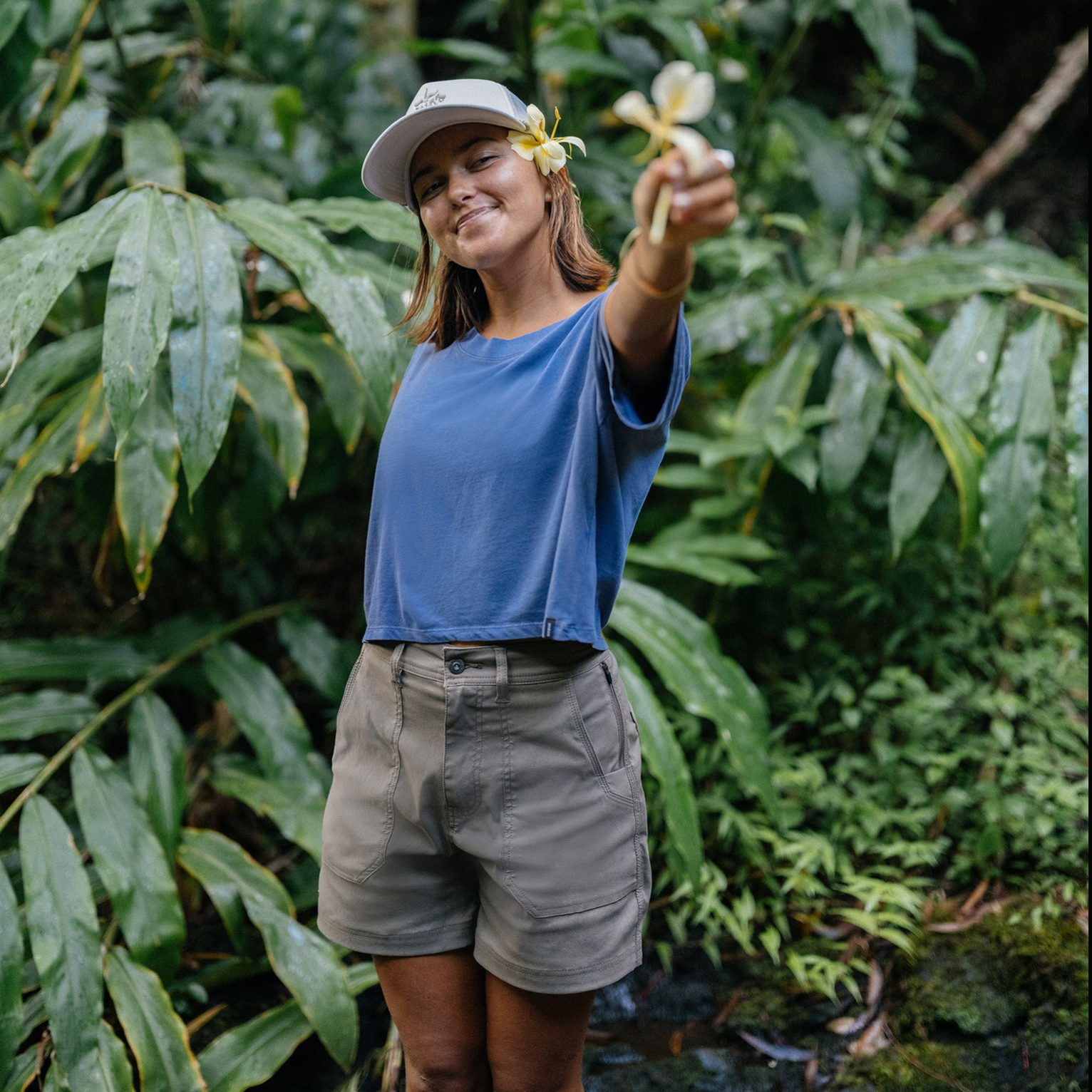 A person in casual outdoor attire, featuring a blue t-shirt, khaki shorts, and a cap, stands in a lush green setting, holding a yellow flower.