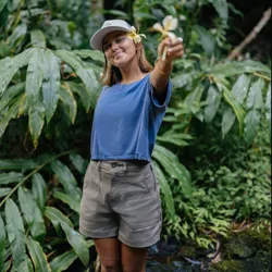 A person in casual outdoor attire, featuring a blue t-shirt, khaki shorts, and a cap, stands in a lush green setting, holding a yellow flower.