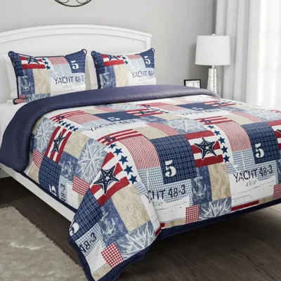A bed features a patchwork quilt and two matching pillow shams with nautical and star designs in red, white, and blue. The room includes a white headboard, bedside table with books, a plant, table lamp, and wall decor above the bed.