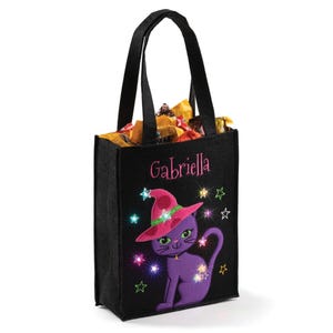 Black tote bag featuring a purple cat wearing a witch hat with stars and personalized with \“Gabriella,\“ filled with assorted candy.