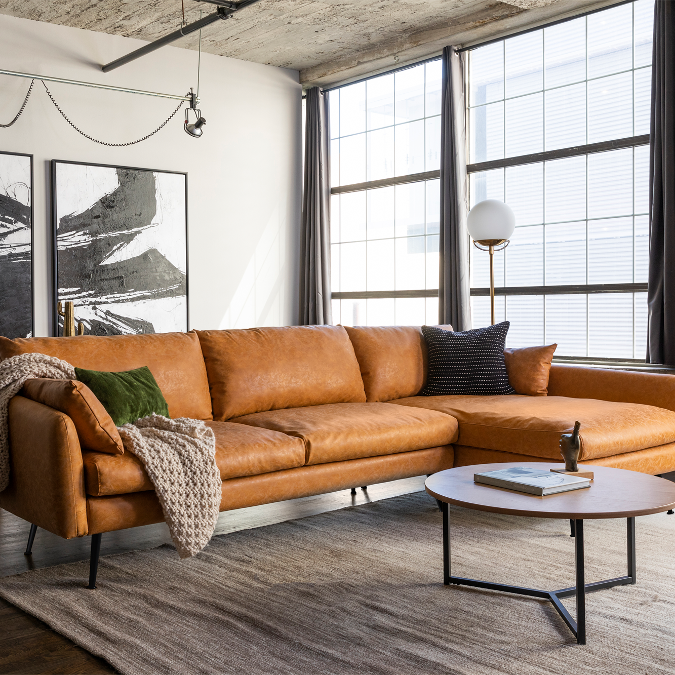 A tan leather sectional sofa with sleek, modern lines features plush cushions and is adorned with textured throws and pillows. It is accompanied by a round coffee table on a large, neutral-colored rug in a bright, spacious living room.