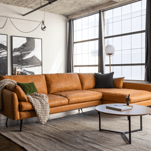A tan leather sectional sofa with sleek, modern lines features plush cushions and is adorned with textured throws and pillows. It is accompanied by a round coffee table on a large, neutral-colored rug in a bright, spacious living room.