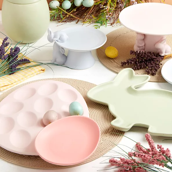 Easter-themed tableware including pastel-colored plates and serving stands, shaped like bunnies and eggs, with decorative elements like artificial flowers and eggs around them.