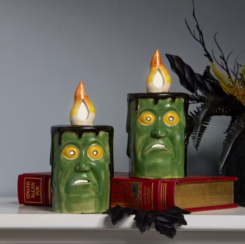 Two ceramic candle holders shaped like green monster faces with flickering light bulbs on top, resting on red books with \“EDGAR ALLAN POE\“ on the spine.