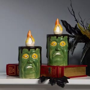 Two ceramic candle holders shaped like green monster faces with flickering light bulbs on top, resting on red books with \“EDGAR ALLAN POE\“ on the spine.