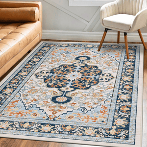 A 5' x 7' machine-washable boho rug features a multicolored, intricate floral pattern with orange and blue hues. It lies on a wooden floor, bordered by a beige sofa and a white chair.