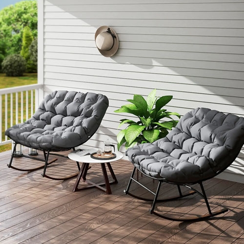 A pair of gray cushioned outdoor rocking chairs with black metal frames, placed on a wooden deck. They are accompanied by a small round table and a potted plant, with a hat hanging on a nearby wall.