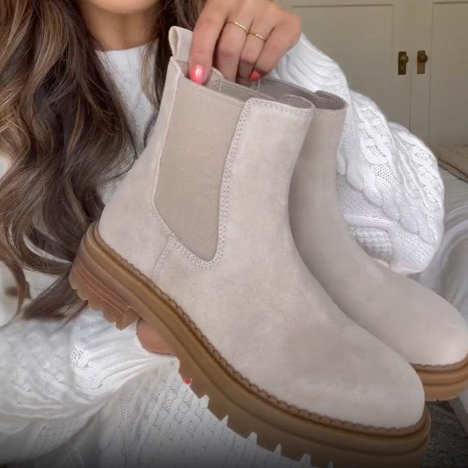 A pair of beige suede Chelsea boots with elastic side panels and ridged soles is being held by a person.