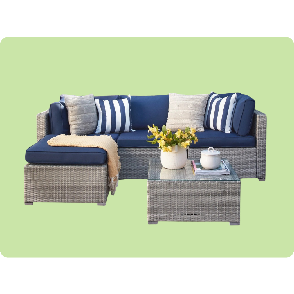 Outdoor patio set with a gray wicker sectional sofa featuring blue cushions and assorted pillows, paired with a matching wicker coffee table topped with a glass surface and decorative items. Background is light green.