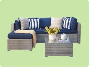 Outdoor patio set with a gray wicker sectional sofa featuring blue cushions and assorted pillows, paired with a matching wicker coffee table topped with a glass surface and decorative items. Background is light green.
