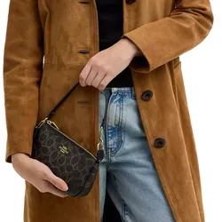 A person is wearing a brown coat and blue jeans while holding a small black Coach wristlet with a gold zipper and logo.