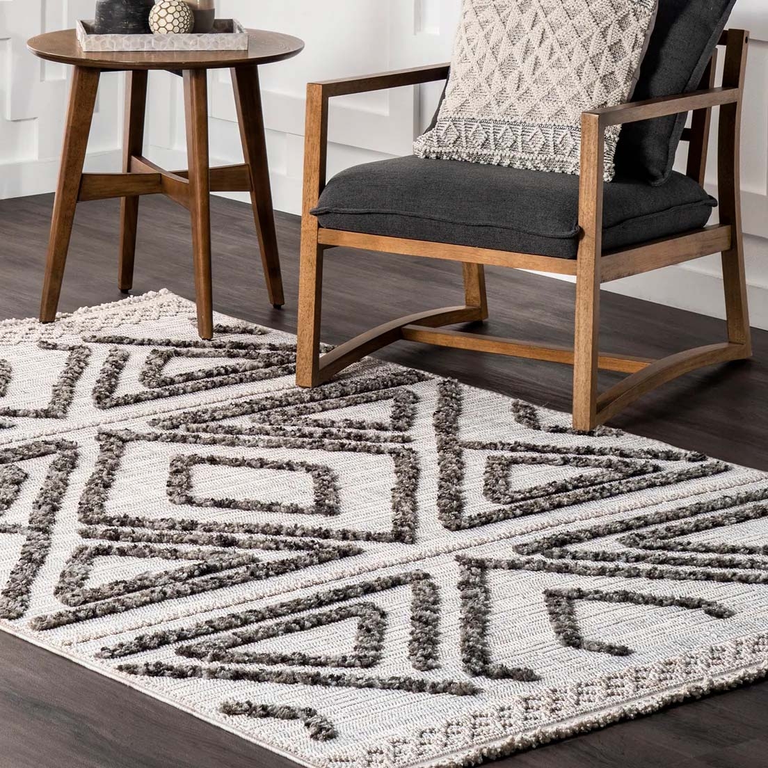 A geometric-patterned rug in neutral tones lies on a dark wood floor, accompanied by a gray cushioned chair with wooden arms and legs and a small round wooden side table holding a tray with decor items.