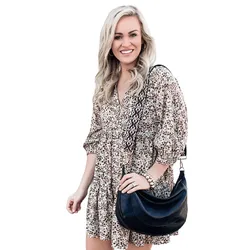 A woman is wearing a floral patterned dress with three-quarter sleeves, holding a black leather shoulder bag with a printed strap. She has long blond hair and is smiling.