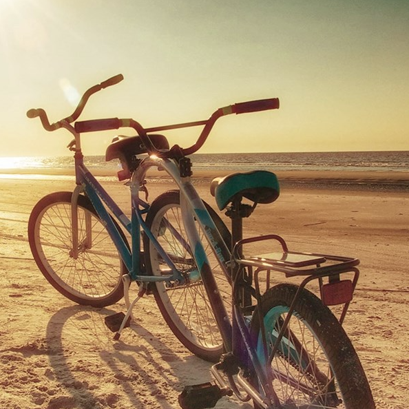 Two bicycles are parked on a sandy beach with the sun setting over the ocean in the background.