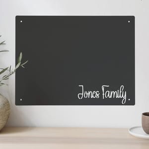 A black rectangular sign with \“Jones Family\“ written in white script is mounted on a wall, next to some greenery and a cup on a wooden surface.