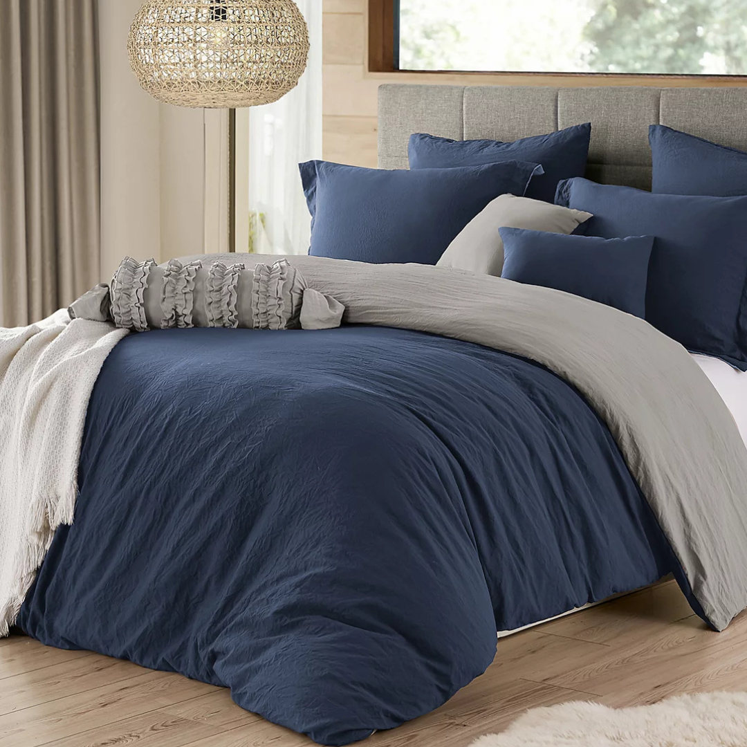 A cozy bedroom setup featuring a navy blue duvet cover, complemented by matching pillows and a light gray throw blanket. The scene includes a textured bolero pillow, a woven pendant light, and a soft area rug on wooden flooring.