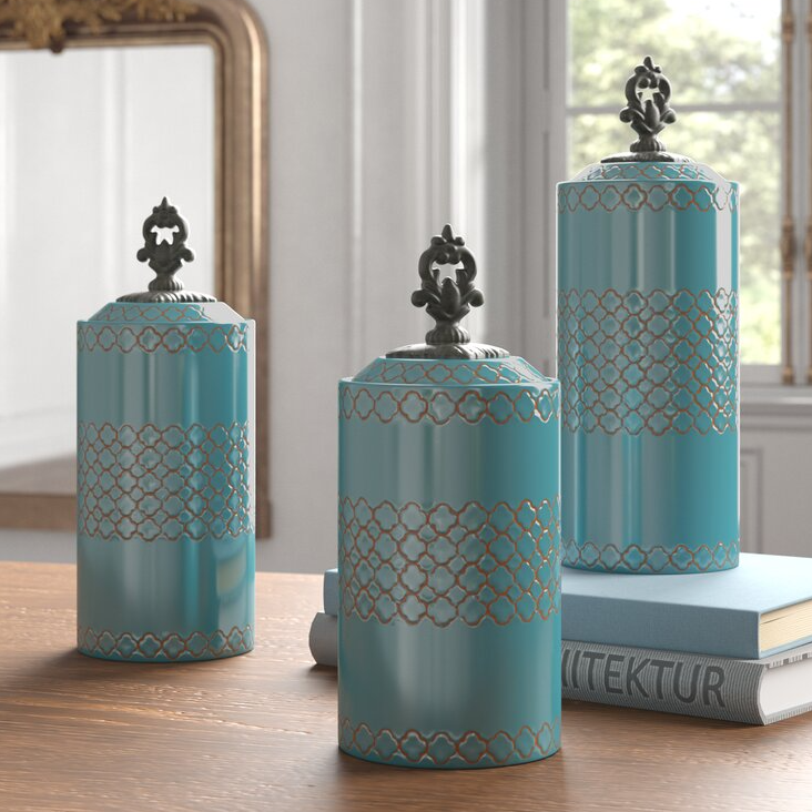 Three turquoise decorative canisters with ornate lids and a lacy pattern, displayed on a wooden surface beside a book.