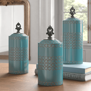 Three turquoise decorative canisters with ornate lids and a lacy pattern, displayed on a wooden surface beside a book.