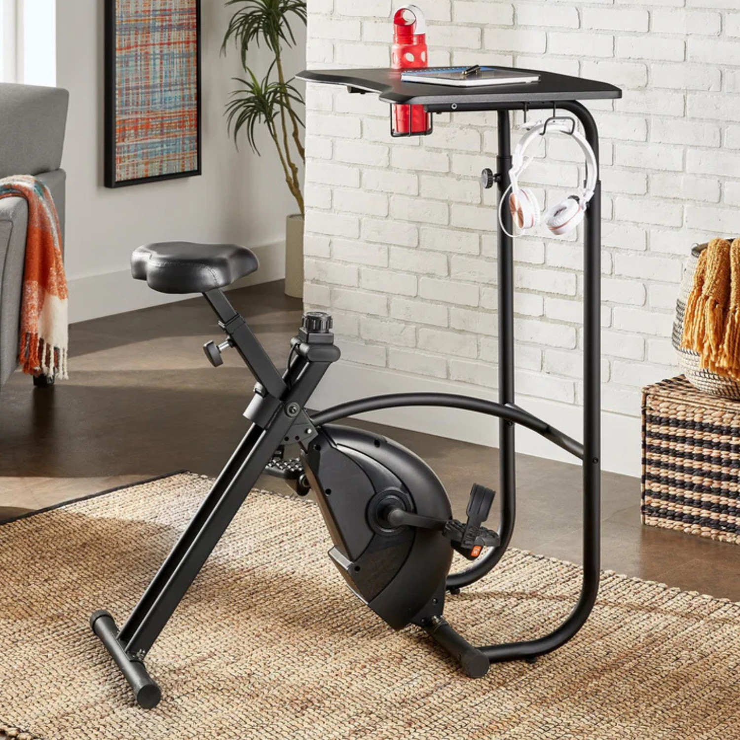 The Virgil Exercise Bike Desk features a stationary bike with an adjustable seat and pedals attached to a built-in desk with space for items, a red water bottle, and headphones. It combines a workout bike and workstation in a space-efficient design.