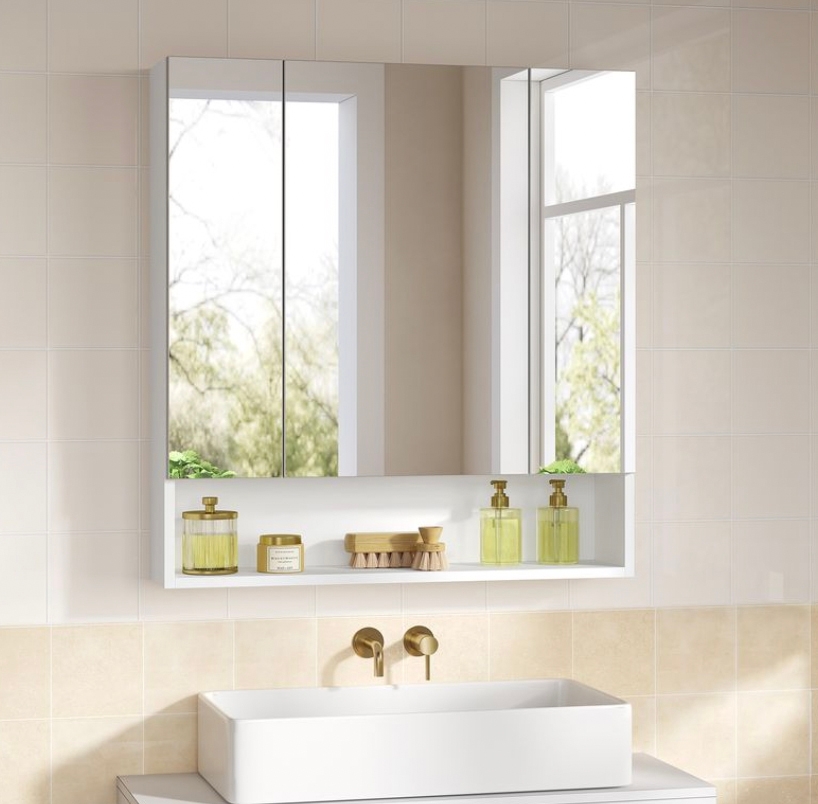 Wall-mounted bathroom mirror with hidden storage, featuring two mirrored doors and an open shelf below. White frame with dual gold dispensers and a small brush placed on the shelf.