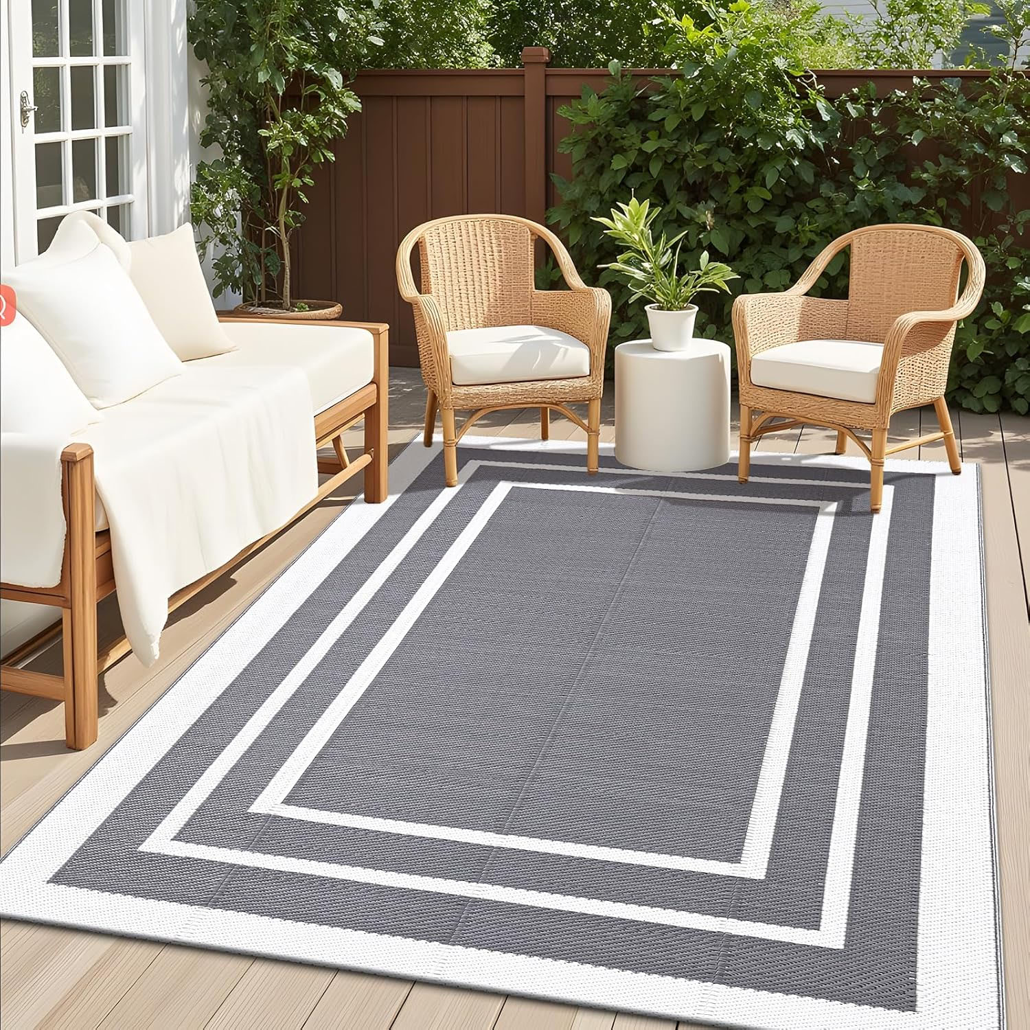 The Olanly 5' x 8' outdoor rug features a gray border design, perfect for patios. It is shown under a seating area with wooden and wicker furniture and surrounded by greenery.