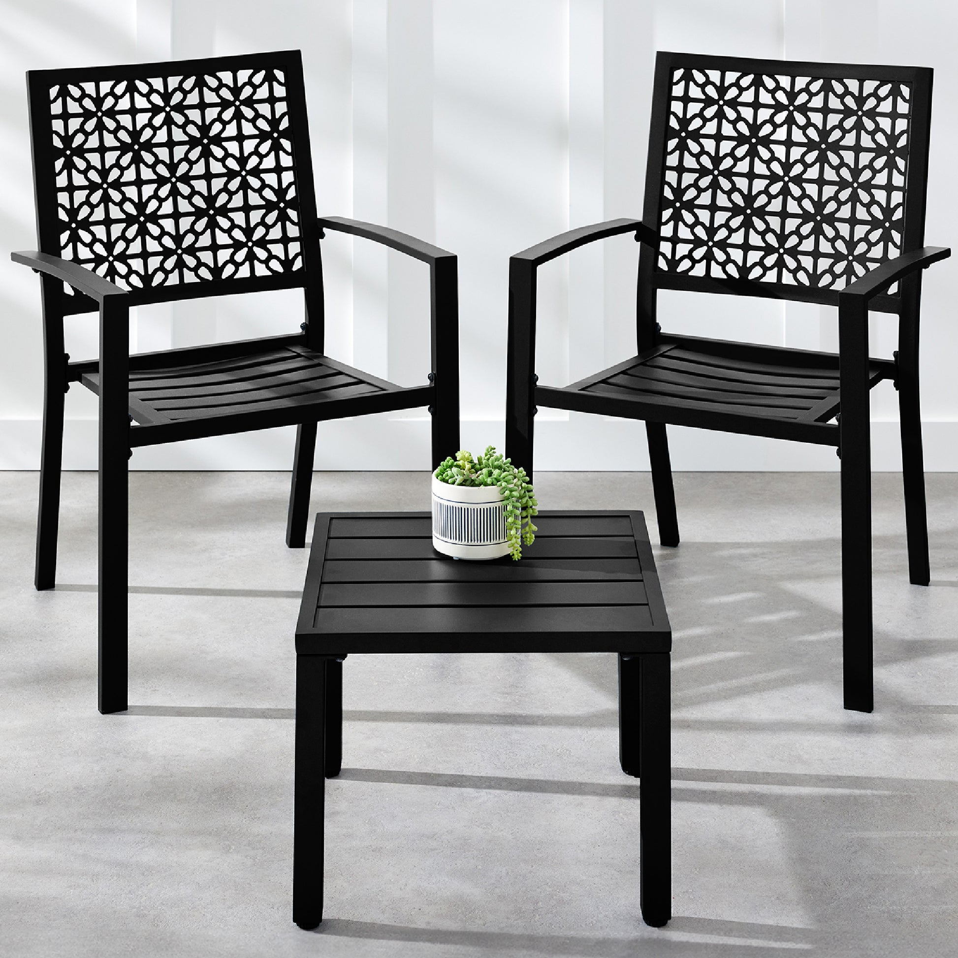 Two black patio chairs with intricate lattice back designs are paired with a matching square side table. A small potted plant is placed on the table.