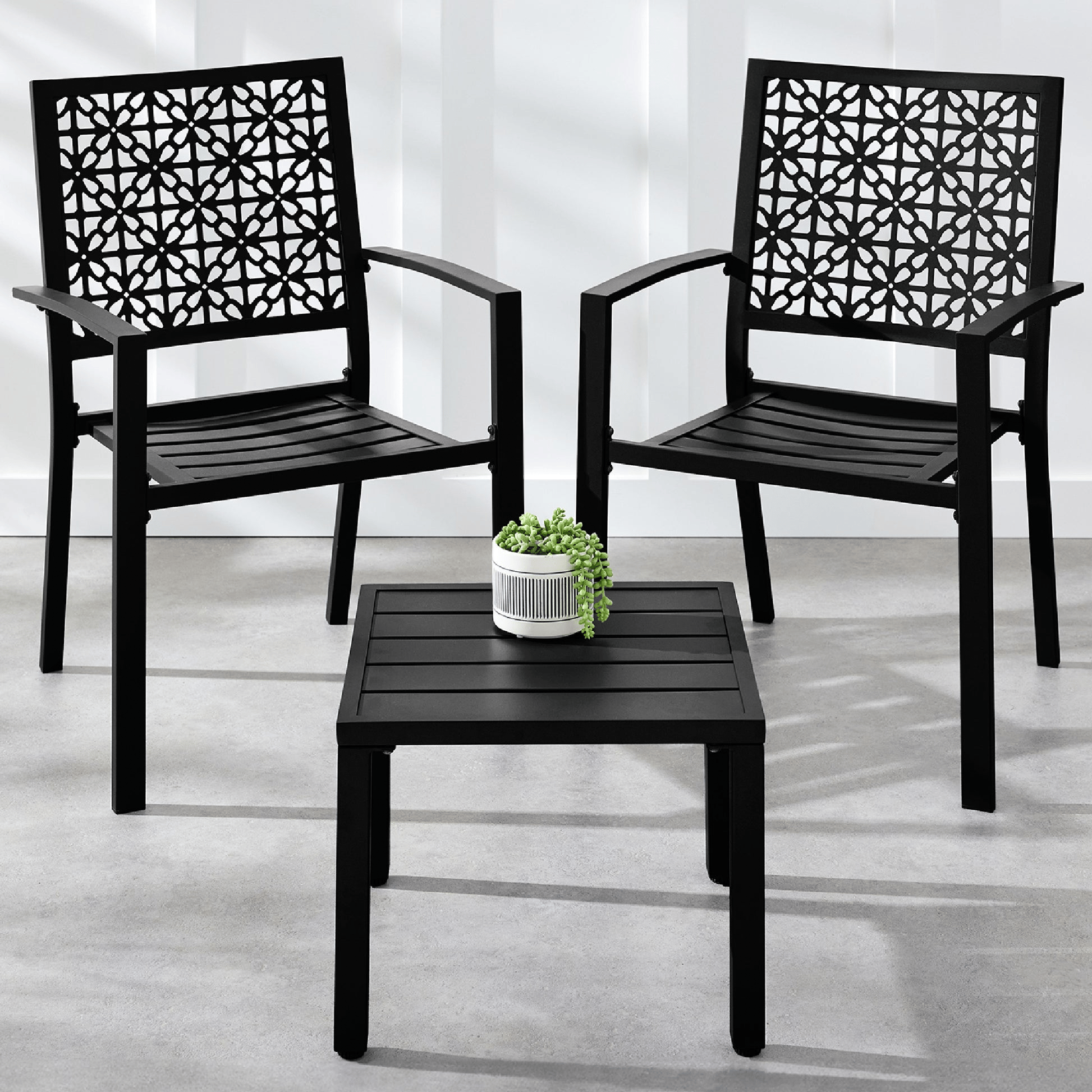 Two black patio chairs with intricate lattice back designs are paired with a matching square side table. A small potted plant is placed on the table.