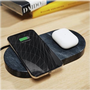 A phone and a set of wireless earbuds are charging on a dual-sectioned stone pad with two separate charging areas, one made of stone and the other covered in fabric.