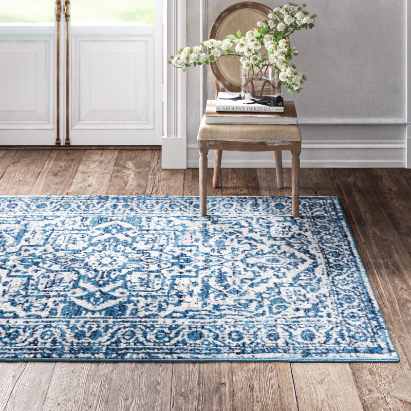 A blue and white patterned rug with a vintage design is on a wooden floor. A wooden chair with a cushioned seat holds a vase with flowers and books, near a white-paneled door.