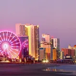 Cityscape featuring a Ferris wheel illuminated at night, set against a backdrop of modern skyscrapers and a coastline.