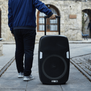 A person is pulling a portable speaker with a handle, resembling luggage, on a city street. The speaker features a large front-facing woofer and carries a visible Altec label.