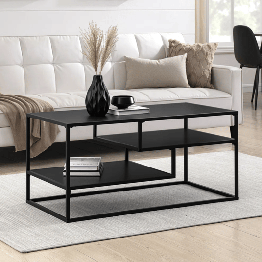 Black metal coffee table with two shelves set on a light gray rug, positioned in front of a white tufted sofa. Decor includes a black vase with pampas grass, a book, a candle, beige and knit pillows, and abstract wall art.