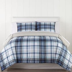 A bed features a plaid-patterned comforter and matching pillow shams in shades of blue, white, and black. The setting includes a neutral backdrop with textured white walls.