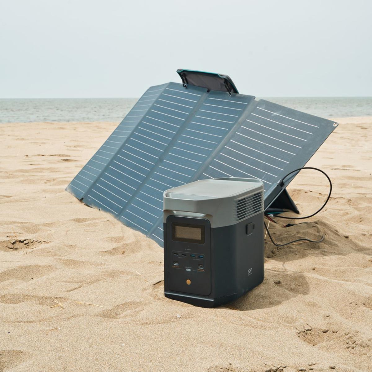 A portable power station with a large display sits on the sand, connected to foldable solar panels propped up to absorb sunlight.