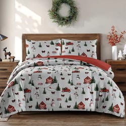 A festive bedding set featuring a winter holiday theme with snow-covered houses, red trucks, trees, and snowmen. It includes two pillowcases on a wooden bed with a wreath and holiday decorations nearby.