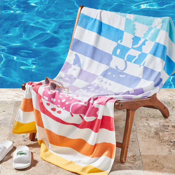 A chair by a pool is draped with a colorful striped towel featuring animal silhouettes. Nearby are white slides with a green logo and a pair of sunglasses.