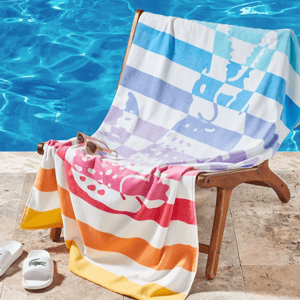 A chair by a pool is draped with a colorful striped towel featuring animal silhouettes. Nearby are white slides with a green logo and a pair of sunglasses.