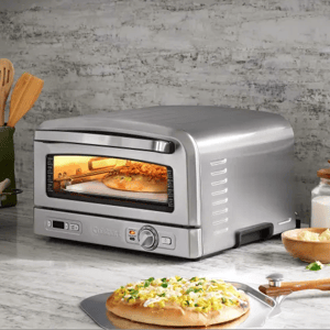 A countertop pizza oven is baking a pizza. The scene includes kitchen utensils, cheese, and a milk bottle on a marble countertop.