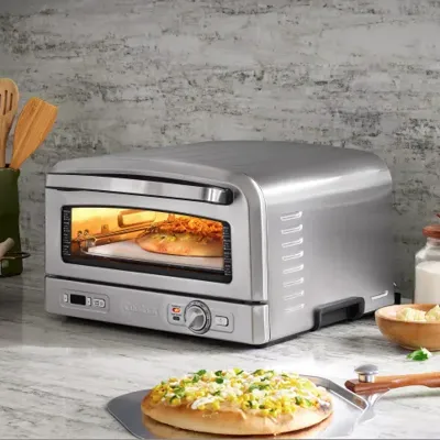 A countertop pizza oven is baking a pizza. The scene includes kitchen utensils, cheese, and a milk bottle on a marble countertop.