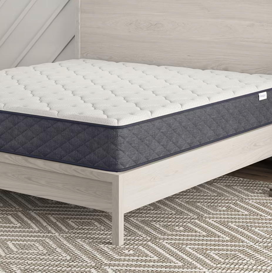 A bed featuring a mattress with a quilted white top and dark blue sides is set in a modern room, accompanied by a decorative round side table and geometric-patterned rug.