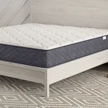 A bed featuring a mattress with a quilted white top and dark blue sides is set in a modern room, accompanied by a decorative round side table and geometric-patterned rug.