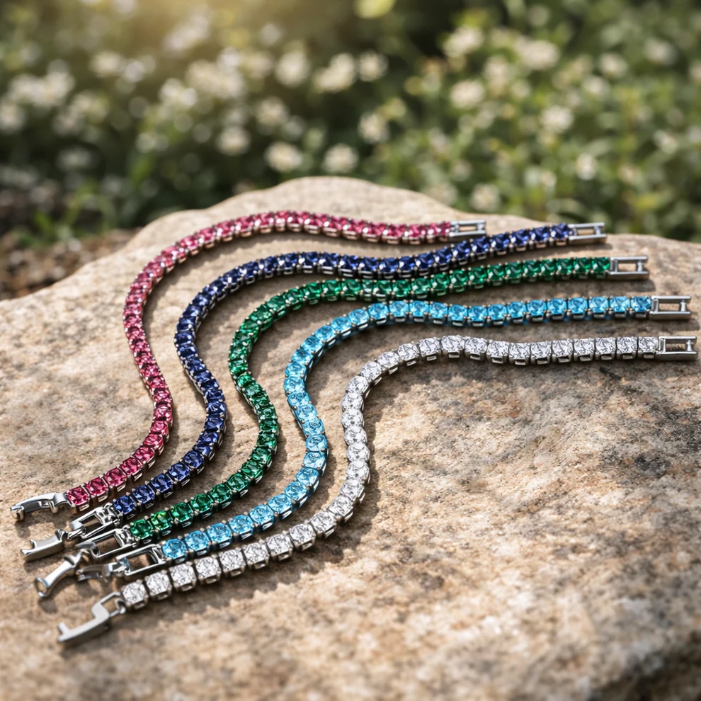 Five tennis bracelets with square-cut stones in pink, blue, green, light blue, and clear colors are displayed on a rock outdoors.