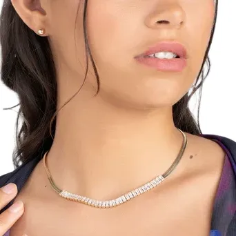 A woman wearing a two-toned necklace with a curved design featuring small stones, along with a small stud earring. Her hair is styled in loose waves.