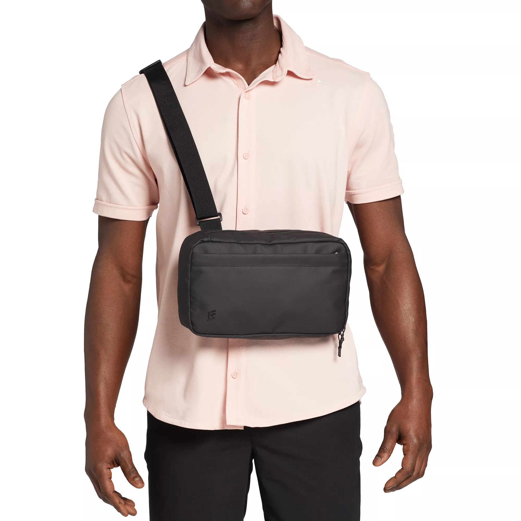 A man is shown wearing a black VRST crossbody bag over his chest, featuring an adjustable strap. He is dressed in a light pink short-sleeve shirt and black pants.