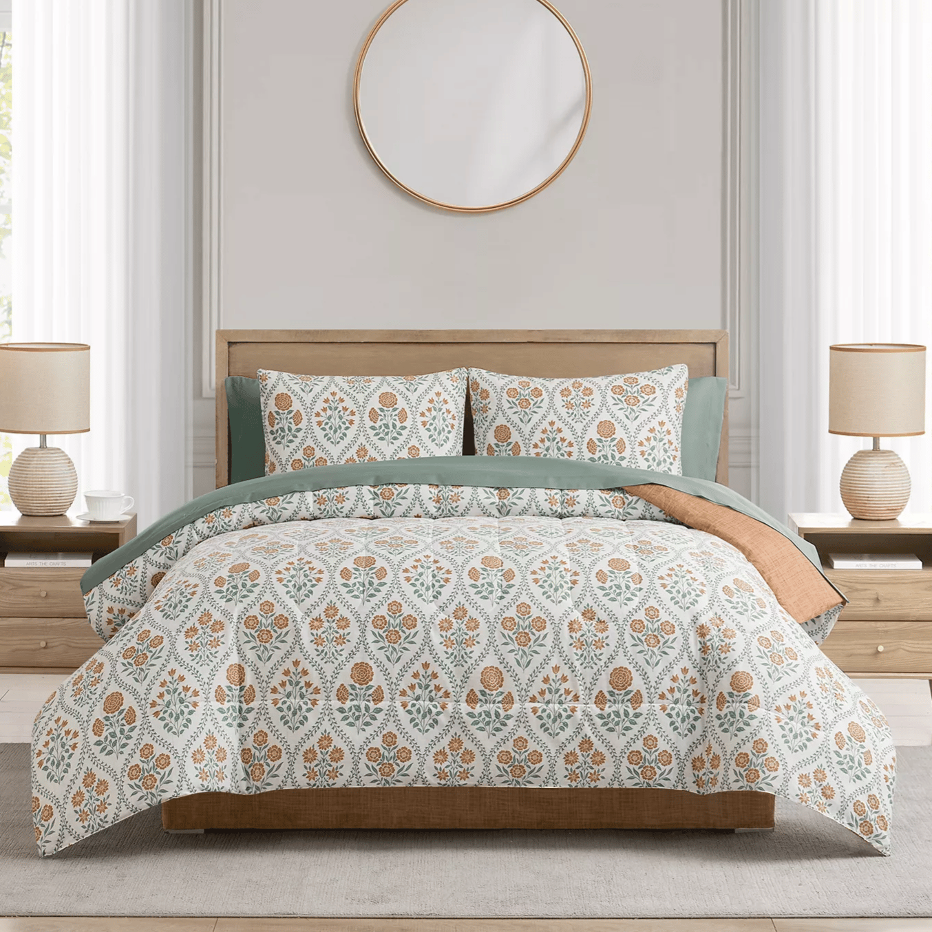 A bed with a floral-patterned comforter set, green and rust-colored accent pillows, wooden headboard, two matching bedside tables with beige lamps, and a circular mirror above completes the bedroom setting.