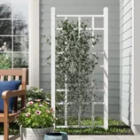 A white outdoor trellis with a grid design supports climbing plants, anchored securely into a small patch of grass, complemented by a nearby wooden bench with a blue cushion and a pot of pink flowers.